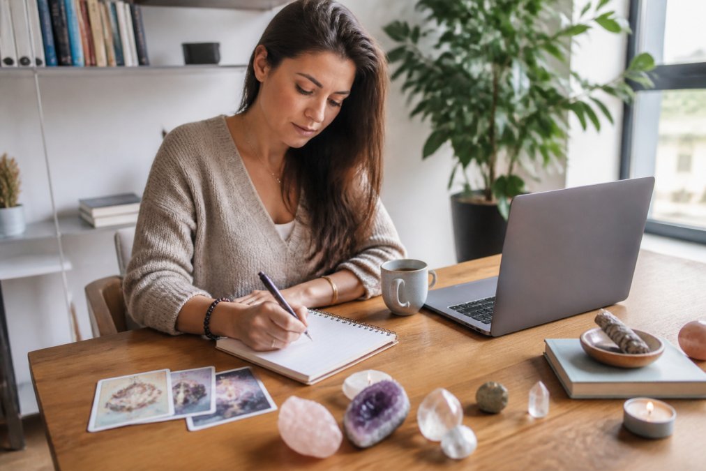 Frau sitzt am modernen Schreibtisch im hellen Büro mit Tarot-Karten, Kristallen und Notizbuch neben ihrem Laptop, arbeitet konzentriert und verbindet dabei spirituelle Praxis mit beruflichem Alltag