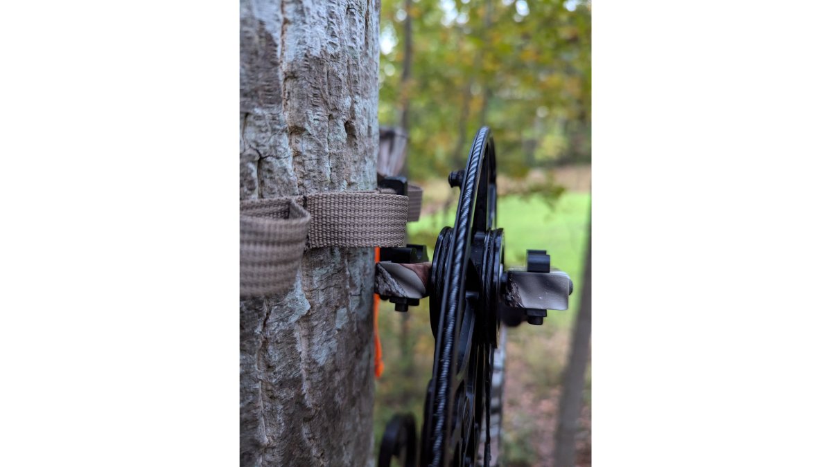 Close-up of a hunter's gloved hand attaching a 3D-printed bow hanger to a gear strap on a tree saddle, compound bow hanging nearby in a forest setting