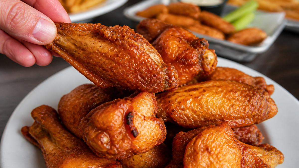 Close-up of freshly cooked golden-brown wings on a white plate with crispy coating and sauce gloss, showing texture detail with a hand holding one wing to demonstrate firmness