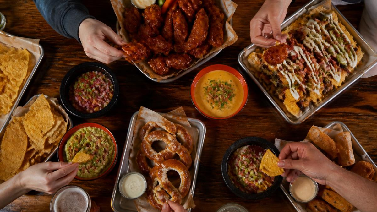 Overhead view of a large wooden table with multiple appetizer platters spread across it, wings glistening with sauce, loaded nachos, warm pretzels, and colorful dips, with hands reaching in to grab food during a game
