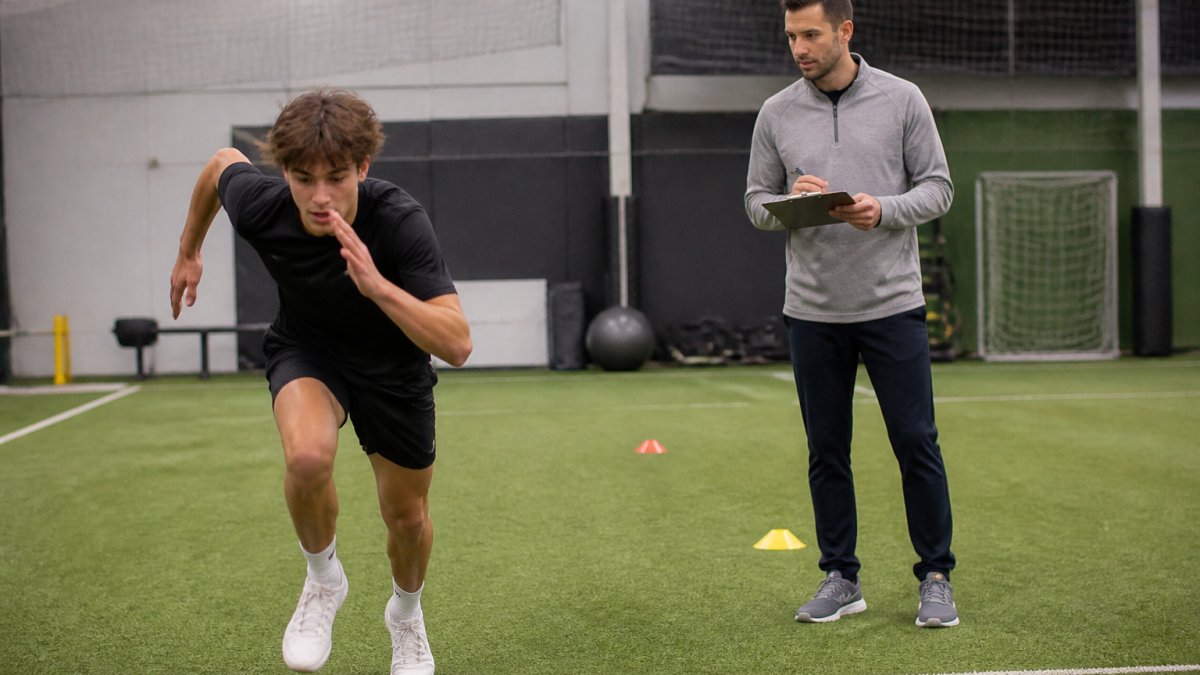 A teenage athlete performing a sprint acceleration drill on an indoor turf field while a physical therapist with a clipboard watches and gives cues