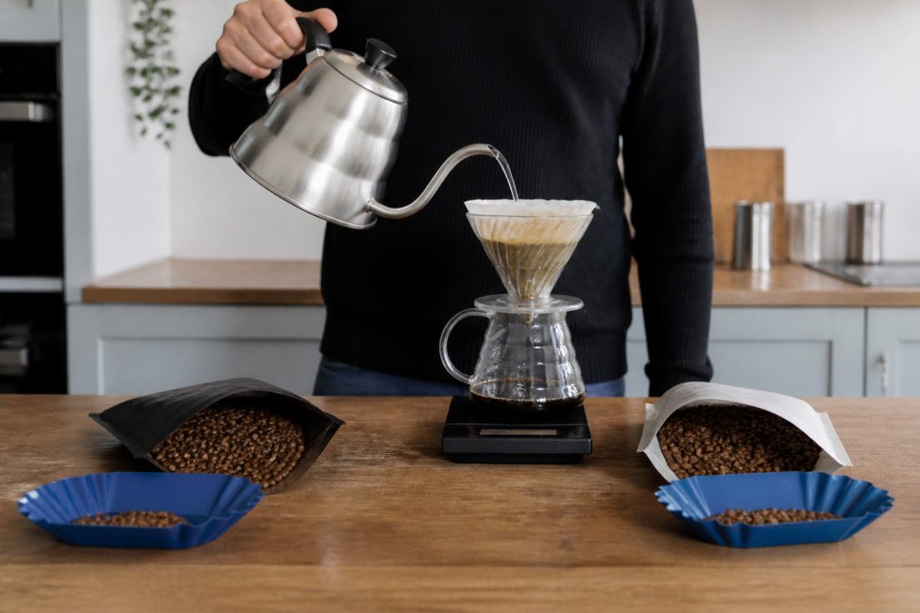A home barista pouring hot water from a gooseneck kettle over a V60 filter, with two open bags of coffee beans and a digital scale on a wooden kitchen counter