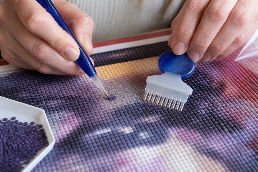 Hands holding a wax pen and multi-placer tool carefully positioning square drills onto canvas grid, showing the precision alignment required and the concentrated focus needed for placement
