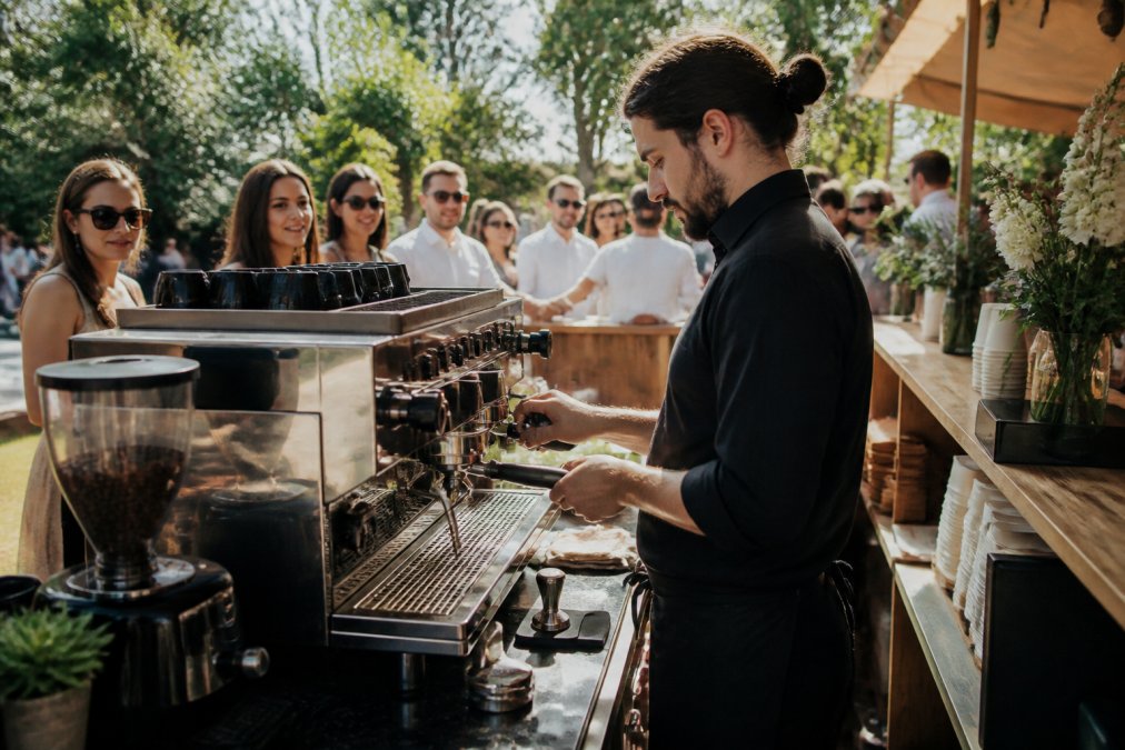 Professional barista operating a La Marzocco espresso machine at an outdoor event, pulling shots with precision while guests watch and wait in line