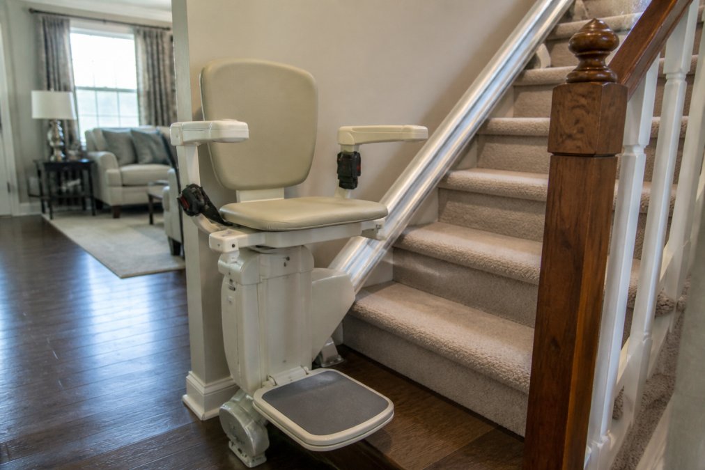 Close-up of an installed stairlift on a residential staircase showing the motorized seat, armrests, safety rails, and track system in a traditional home setting with natural light