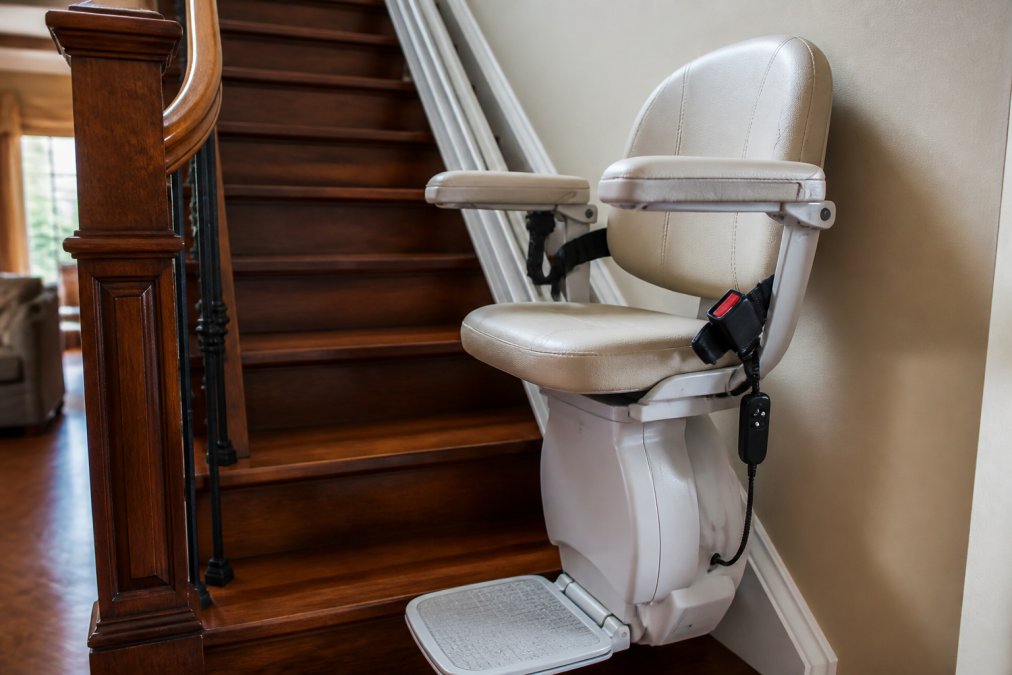 Close-up of an installed stairlift seat showing safety features including padded armrests, seat belt attachment, footrest, and handheld remote control on a residential wooden staircase with natural afternoon light