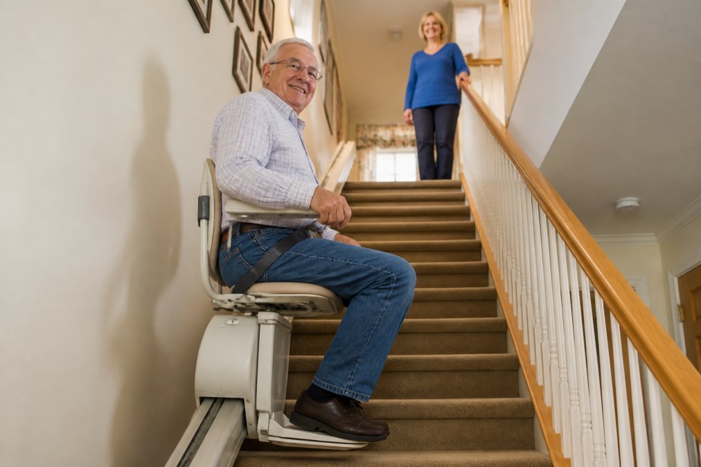 An older adult seated comfortably on a stairlift chair, riding up a carpeted residential staircase with a family member watching supportively from the top landing, warm indoor lighting