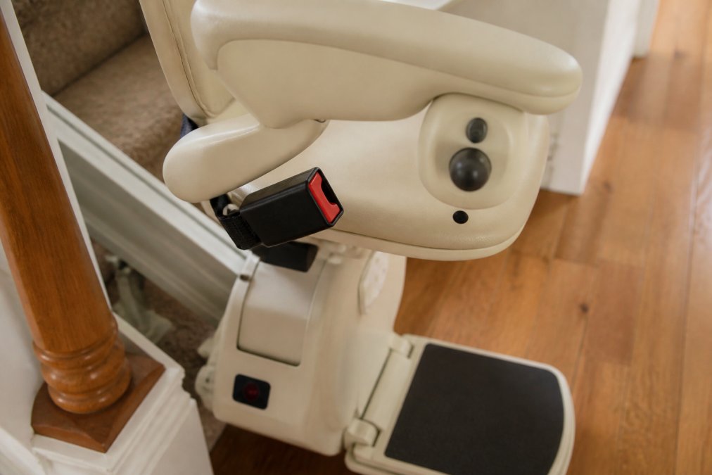 Close-up of a stairlift seat belt buckle and armrest controls showing the swivel seat mechanism and safety sensor panel on a modern stairlift unit, bright indoor lighting