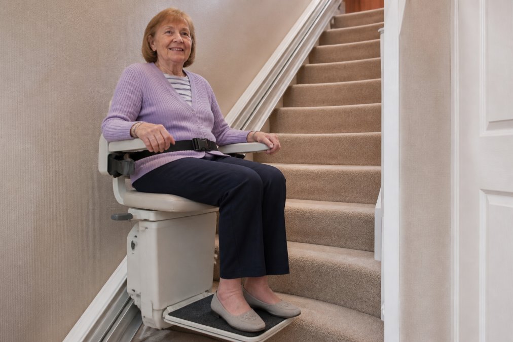 An older adult securely fastened in a stairlift seat with visible seatbelt across the lap, armrests positioned at both sides, and footrest extended, demonstrating proper seating position and restraint