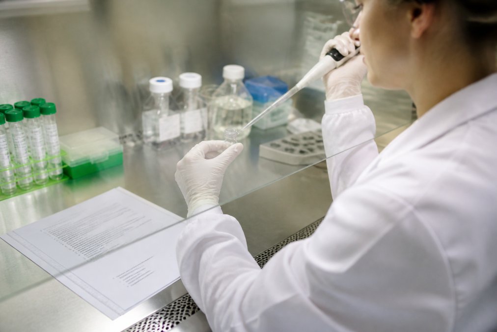 A researcher in a white lab coat and gloves working at a clean bench, carefully handling a small glass vial and a pipette beside a printed standard operating procedure document, with laboratory glassware softly out of focus in the background