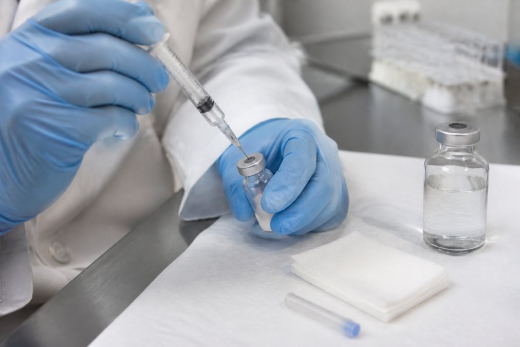 Close-up van een laboratoriummedewerker met handschoenen die een injectiespuit met naald in een injectieflacon met rubberen stop plaatst, op een schone werkbank met steriele doekjes en een flesje bacteriostatisch water ernaast