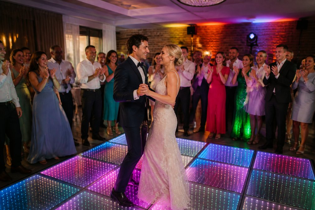 Bruidspaar danst op een verlichte dansvloer met RGB-verlichting tijdens hun bruiloftsfeest, gasten dansen en klappen op de achtergrond in een sfeervol verlichte feestzaal