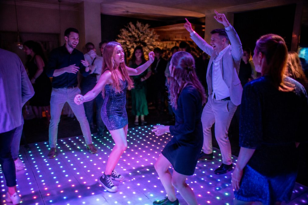 Groep feestgangers dansend op een verlichte dansvloer met kleurrijke RGB-effecten, grote glimlachen, feestelijke sfeer in een zaal met andere gasten op de achtergrond