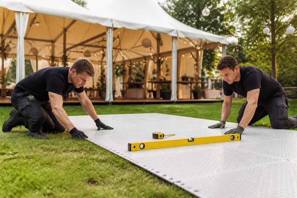 Twee personen leggen kunststof onderlaag neer op gras voor dansvloer-installatie, waterpas en meetlint zichtbaar, voorbereiding van buitenevenement, zorgvuldigheid op gezichten