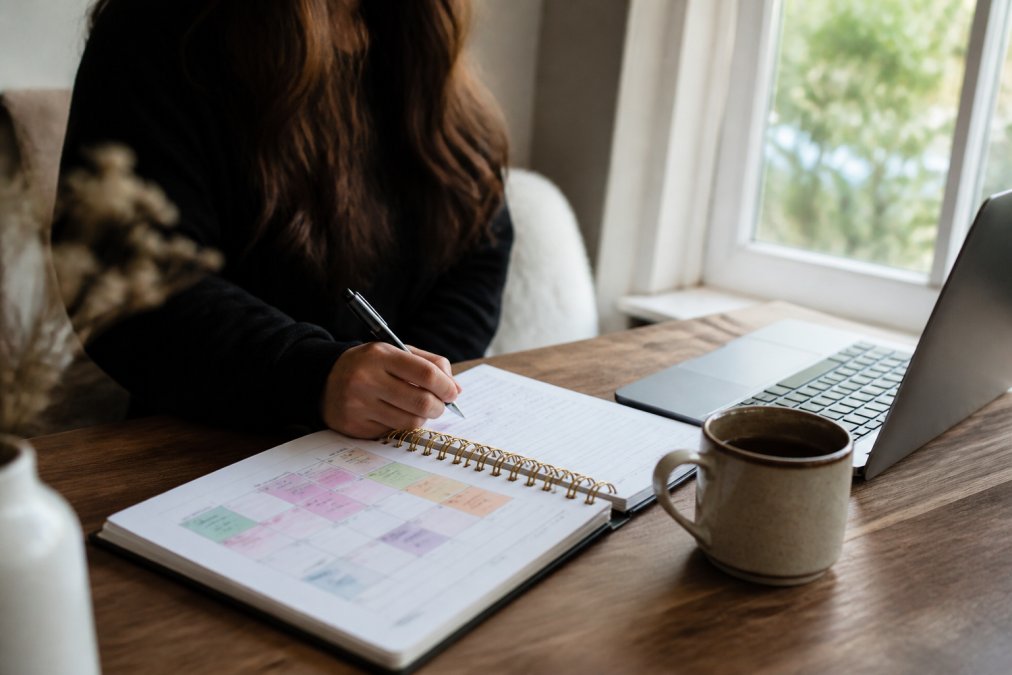 Person working at a laptop at home with a notebook displaying a content calendar with color-coded content pillars and planning notes, a coffee cup nearby, and bright natural window lighting