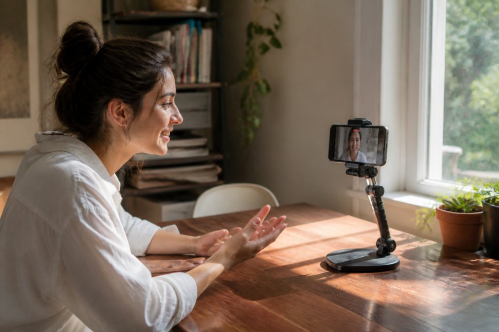Content creator recording video using natural window light and smartphone mounted on adjustable desktop stand in home setup, soft daylight streaming across work surface