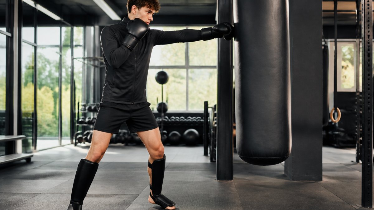 Beginner kickbokser in trainingspak met Booster Alpha handschoenen en scheenbeschermers die een stoot oefent op een bokszak in een moderne sportschool met natuurlijk licht