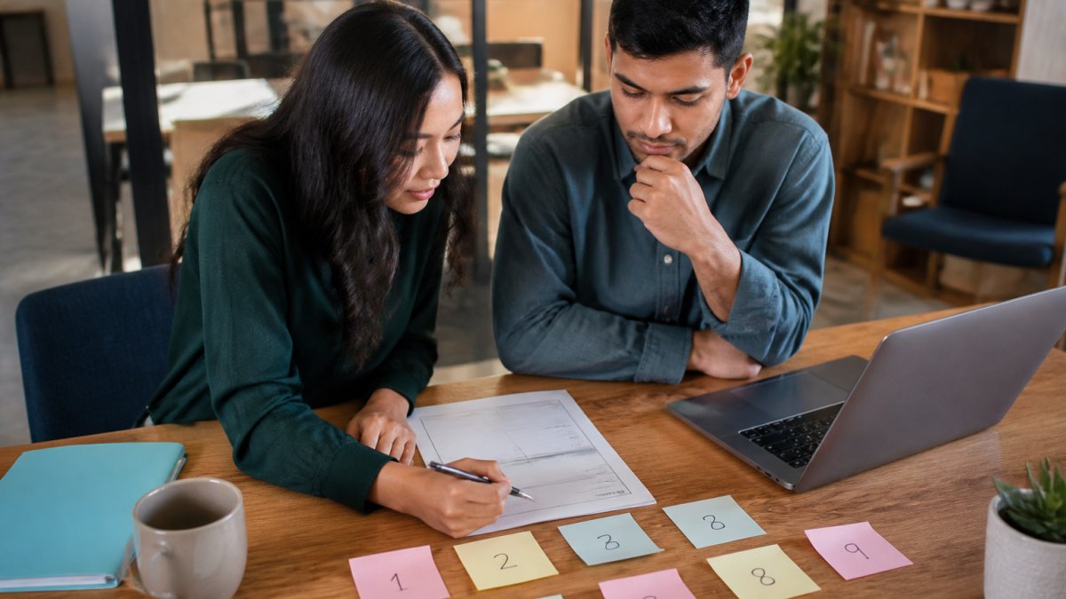 Two founders at a wooden table filling in a printed one-page strategy worksheet with a pen, laptop open beside it, sticky notes with numbers arranged on the tabletop