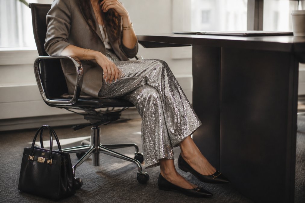 Close-up of professional woman's lower half wearing sequin-embellished trousers in silver, paired with a neutral tailored blazer, delicate gold jewellery, structured leather handbag, and pointed-toe black flats, seated at modern office desk with soft natural lighting