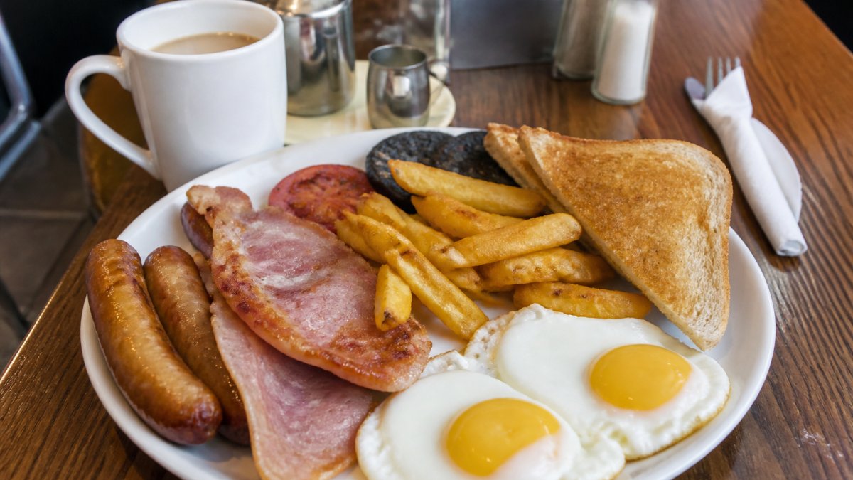 A close-up of a full English breakfast plate with eggs, bacon, sausages, chips and toast on a cafe table, with a mug of coffee beside it