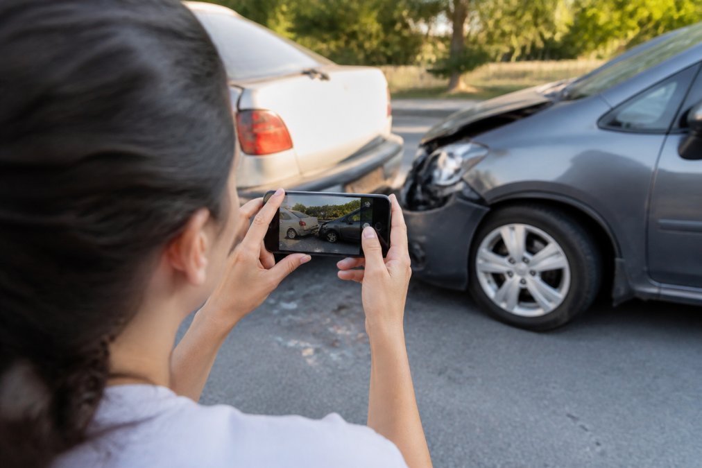 Person photographing visible damage at an accident scene with a smartphone, documenting the scene from multiple angles on a bright afternoon