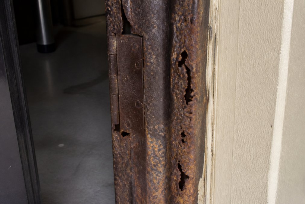 Close-up of a rusted steel door frame showing corrosion and deterioration on the jamb and hinge side, with visible pitting and weathering patterns