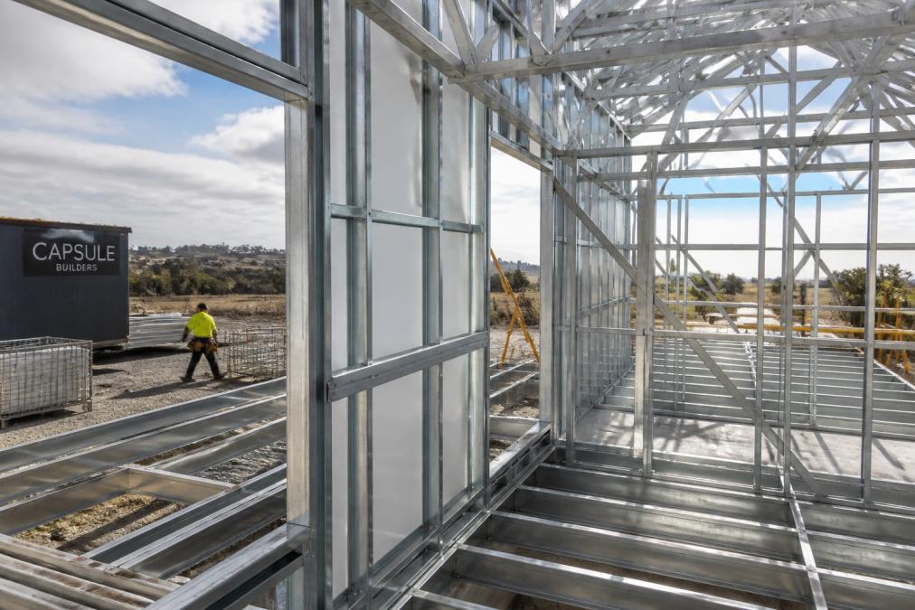 A close-up photograph of a galvanised steel house frame under construction, showing clean, straight lines and precision engineering, with a building site visible in the background