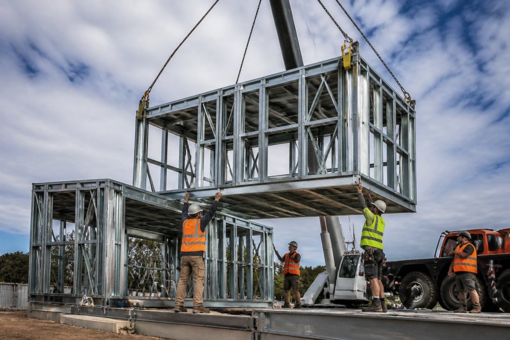 Wide-angle view of a steel-framed modular home under construction, showing prefabricated steel frame modules being assembled on-site with workers in high-visibility gear securing connections