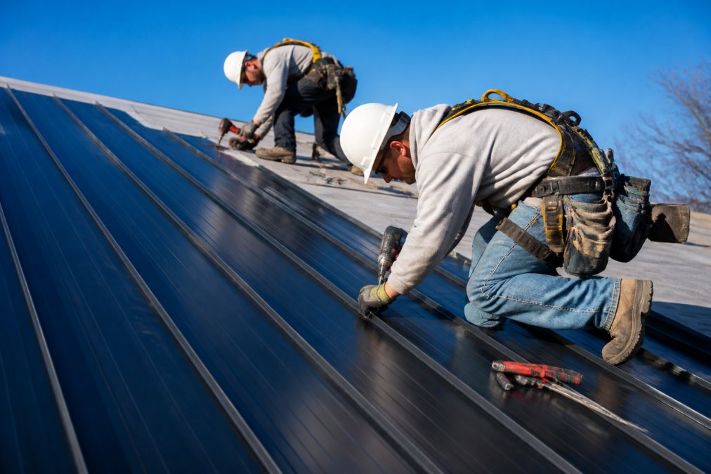 Roofing crew in safety harnesses installing standing seam steel roof panels on a residential home.