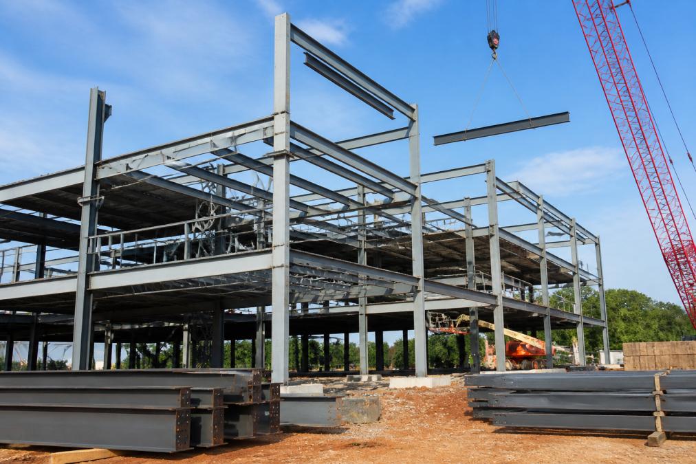 Construction crew installing wide flange steel beams on a multi-story commercial building framework with welding equipment and safety harnesses visible