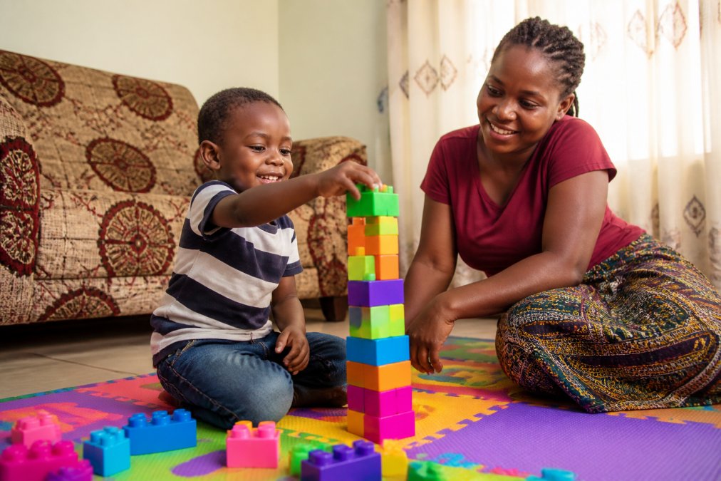 A young child in a bright Nigerian home, smiling while stacking colourful building blocks on a mat, with a parent watching nearby in soft afternoon light