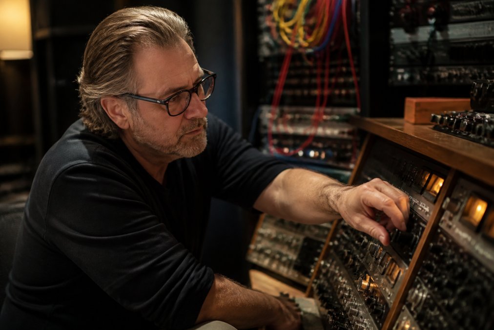 A mastering engineer in a professional studio adjusting analog outboard gear on a console, warm lighting, focused expression, cables and rack units visible in the background