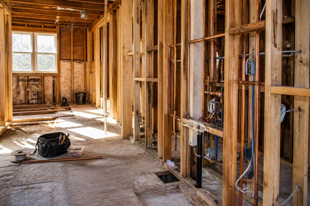 A kitchen mid-renovation with exposed wall studs, new copper plumbing lines and electrical wiring visible, subfloor exposed, tools and construction materials nearby, natural light from a window