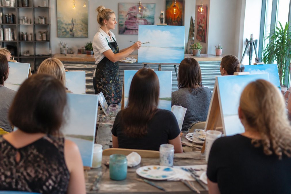 An art instructor demonstrating brush techniques on a canvas at the front of a bright paint and sip studio, with adult students at easels following along in the background, paint palettes and water cups visible on tables