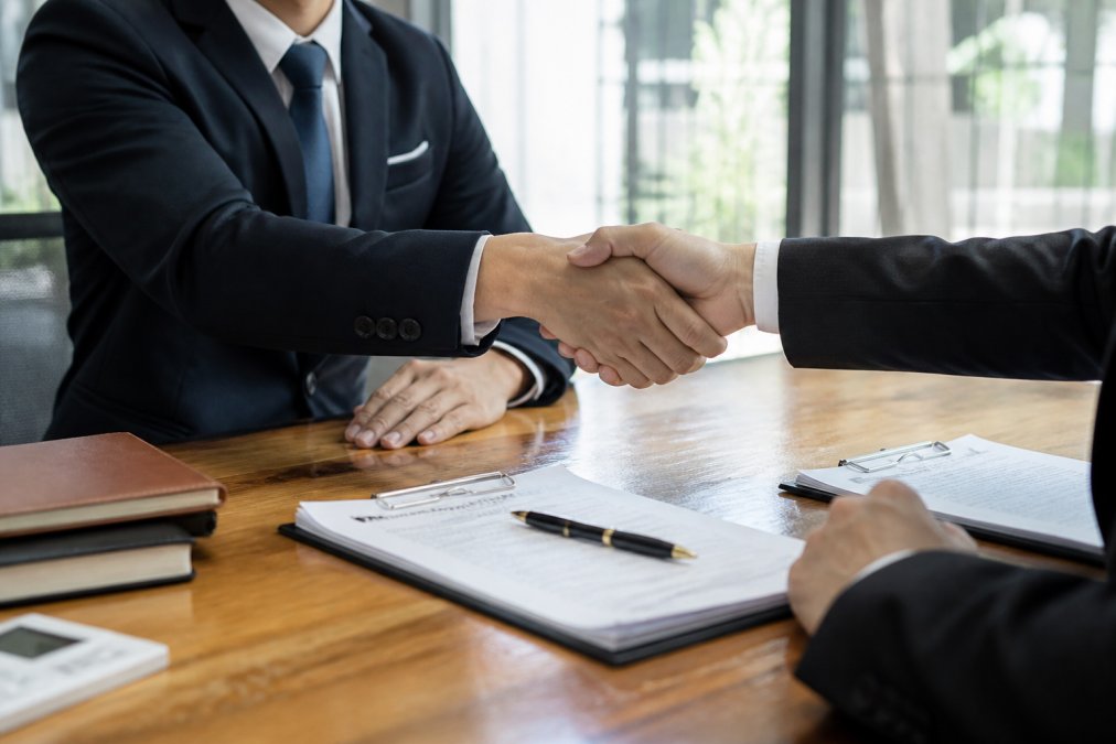 Two business professionals in suits shaking hands across a wooden conference table with a signed contract, pen, and documents visible, indicating agreement on terms