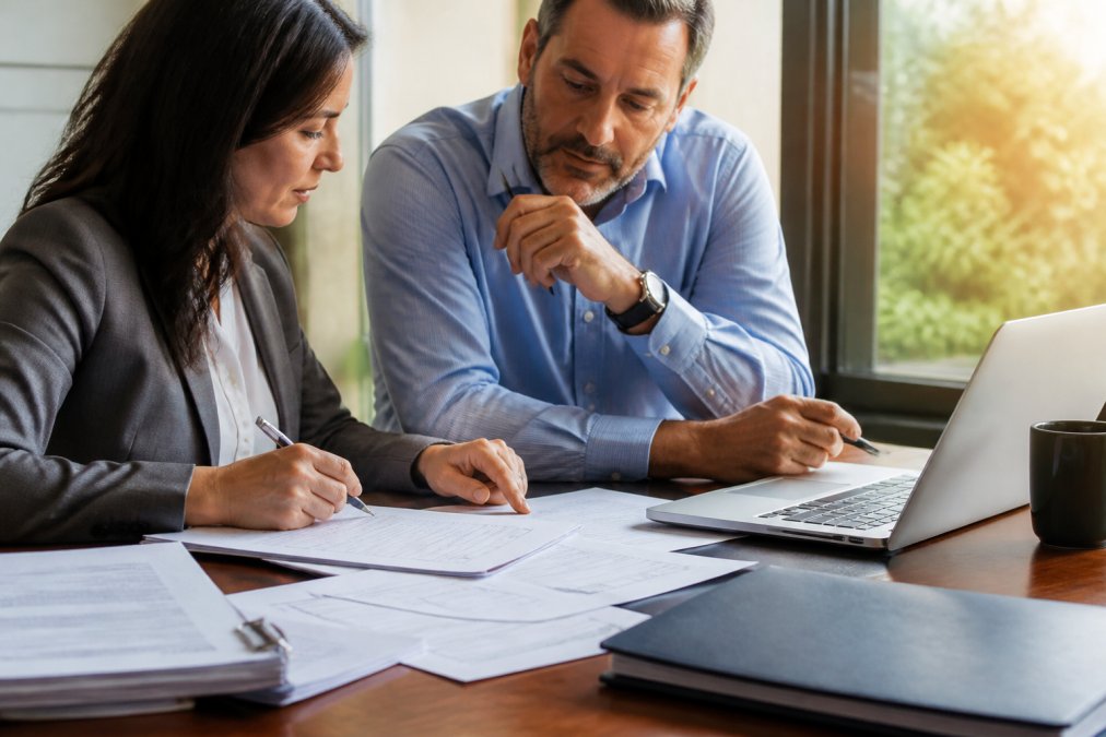 A healthcare consultant and agency owner reviewing licensing paperwork and a checklist at a tidy desk, laptop open to a state health department portal, warm afternoon light