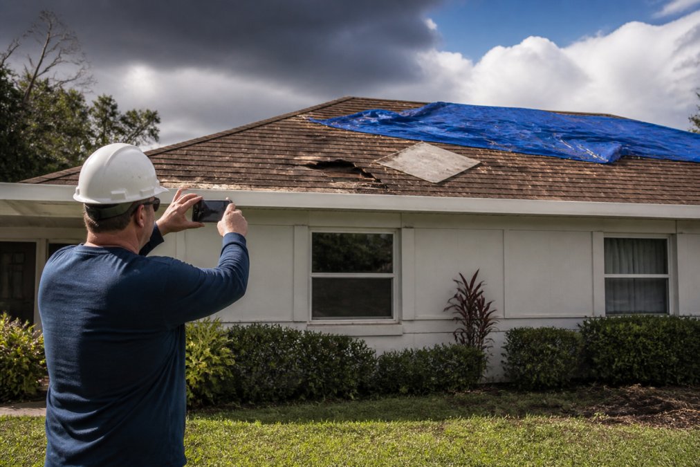 Homeowner in safety gear photographing hurricane roof damage with a smartphone