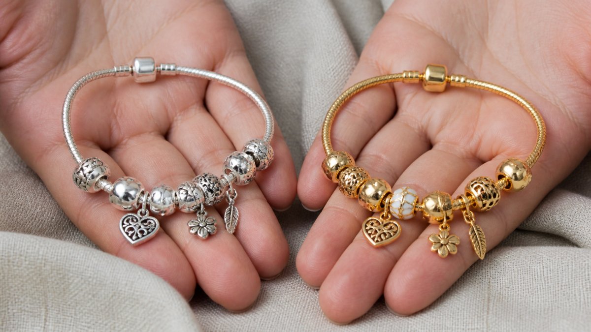 A close-up of two hands holding a sterling silver charm bracelet and a gold plated charm bracelet side by side over a soft neutral fabric surface, natural window light, showing the difference in tone and finish