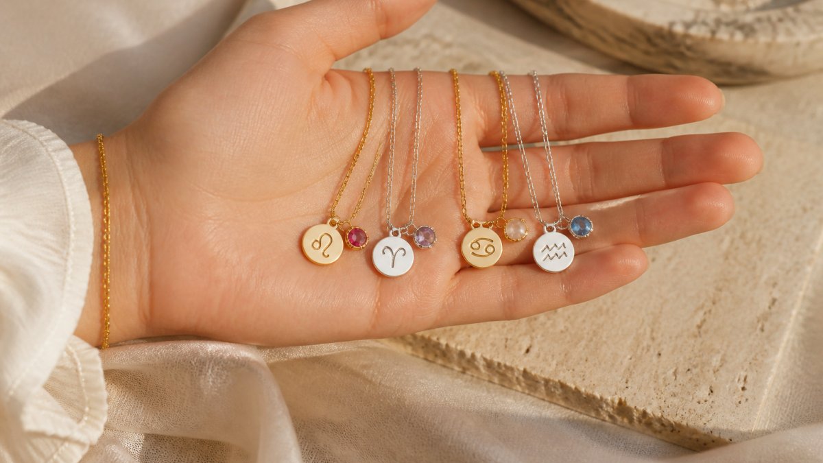 Eine Hand hält mehrere elegante Halsketten mit unterschiedlichen Anhängern (Sternzeichen-Symbole und Edelsteine) nebeneinander, um verschiedene Kombinationsmöglichkeiten zu zeigen. Warmes Licht auf neutralem Untergrund.