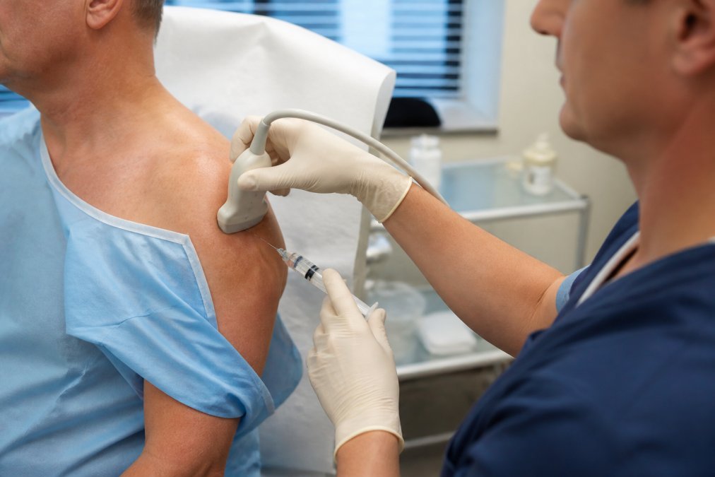 Healthcare professional performing ultrasound-guided shoulder injection procedure on patient in clinical setting, with ultrasound probe and needle visible, patient in examination gown with arm positioned for access