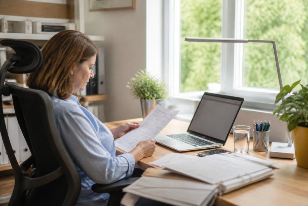 Person sitzt am ergonomischen Schreibtisch mit Laptop und hält Steuerunterlagen in der Hand, während sie auf dem Bildschirm ihre Online-Steuererklärung überprüft, mit natürlicher Fensterbeleuchtung im Hintergrund