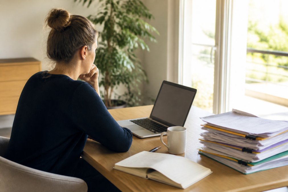 Eine Person sitzt an einem aufgeräumten Schreibtisch mit Laptop, Kaffeetasse und einem Stapel sortierter Dokumente, konzentriert auf den Bildschirm blickend, helles Tageslicht
