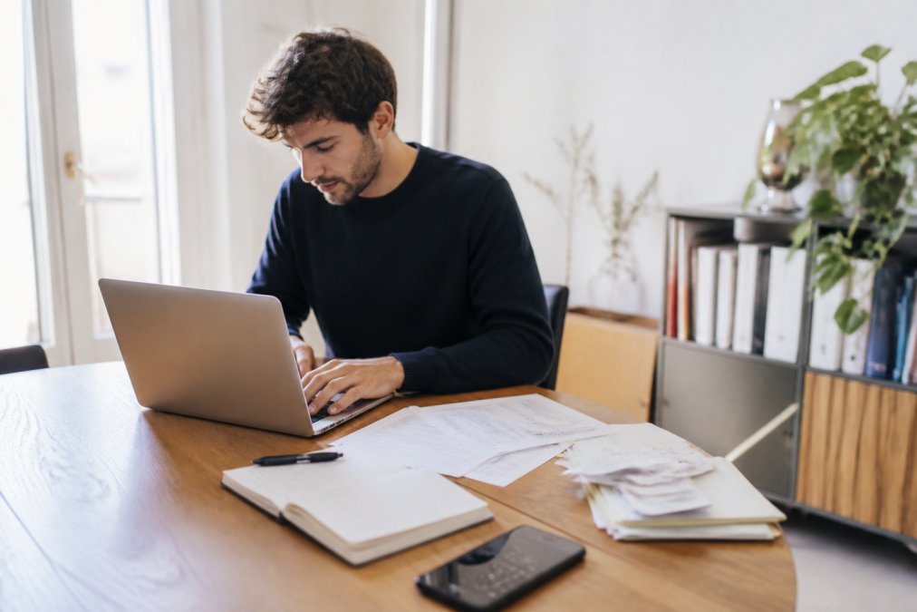 Person sitzt konzentriert an einem Laptop und füllt ein Online-Steuerformular aus, mit Dokumenten, Notizen und Belegen auf dem Schreibtisch daneben, natürliches Tageslicht von der Seite