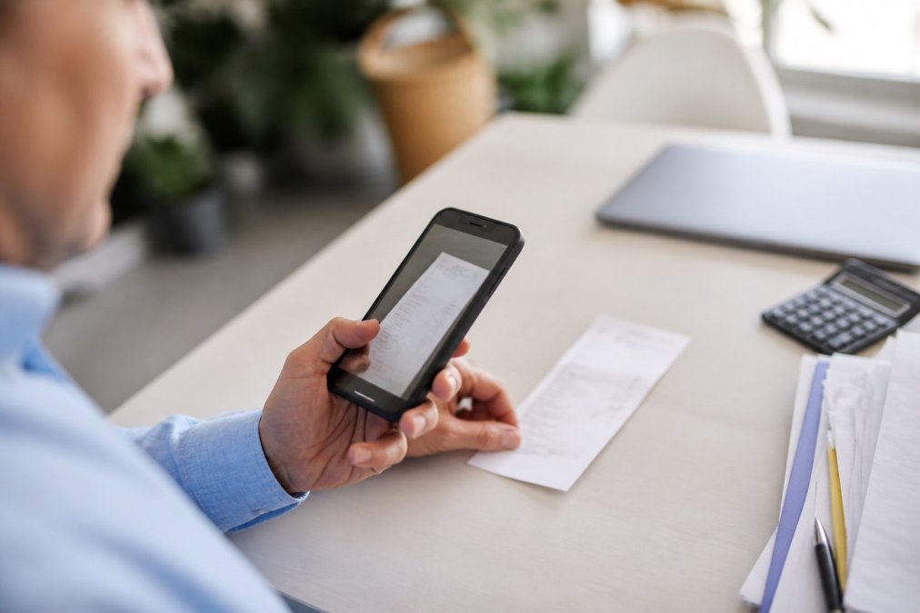 Nahaufnahme einer Hand, die mit einem modernen Smartphone einen Beleg fotografiert, während dieser auf einem hellen Schreibtisch liegt, bereit zum automatischen Upload