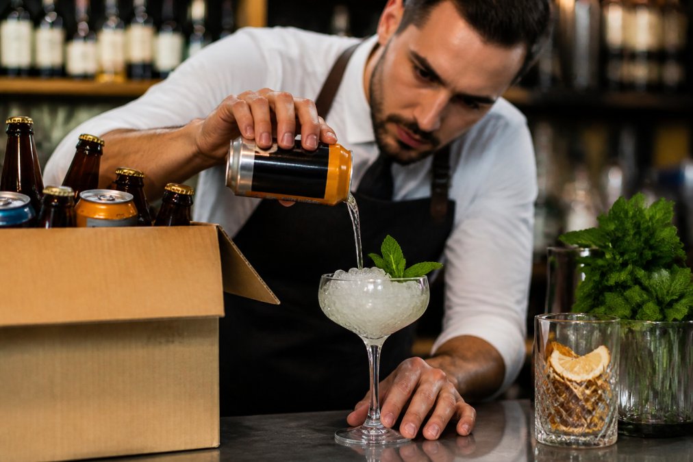 Bartender carefully pouring premium mixer into crystal coupe glass with ice and fresh mint, focusing intently on pour technique with bottles and garnishes arranged on bar counter
