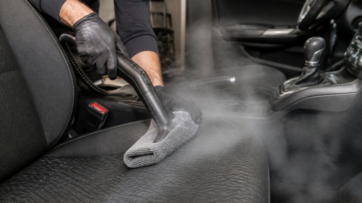Close-up van handen die een stoomreiniger met een microvezeldoek over een autostoel met stoffen bekleding halen, stoomwolk zichtbaar, auto-interieur op de achtergrond