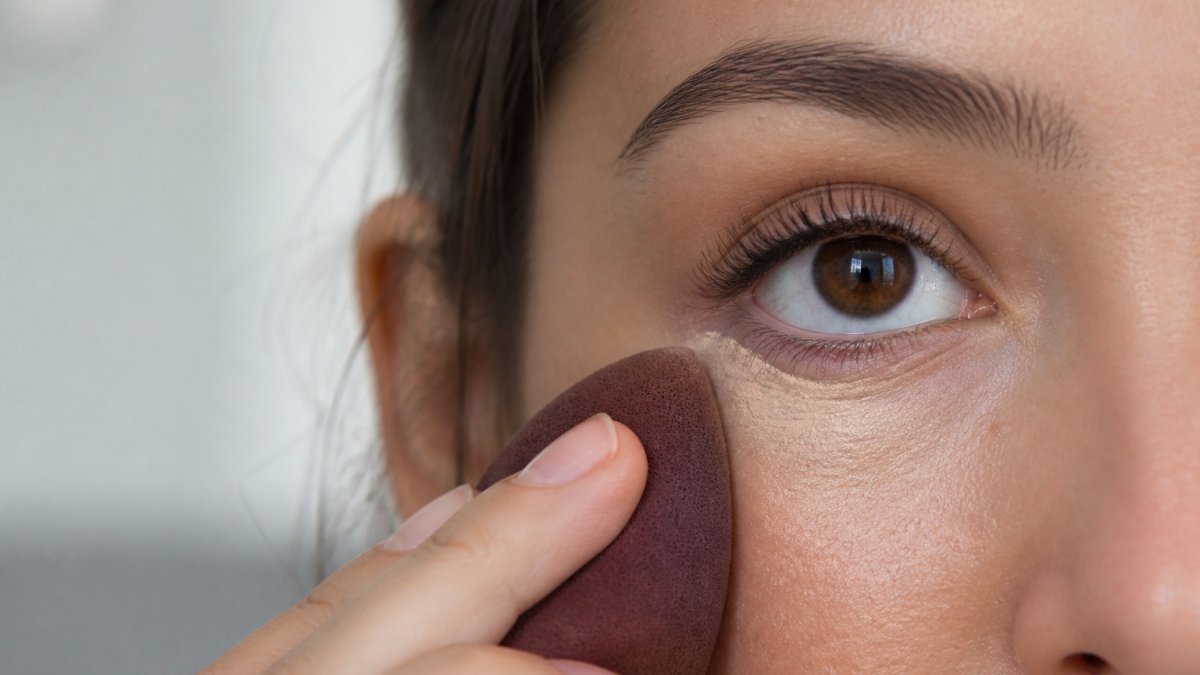 Step-by-step diagram showing a damp sponge gently blending concealer into the under-eye area to prevent creasing.
