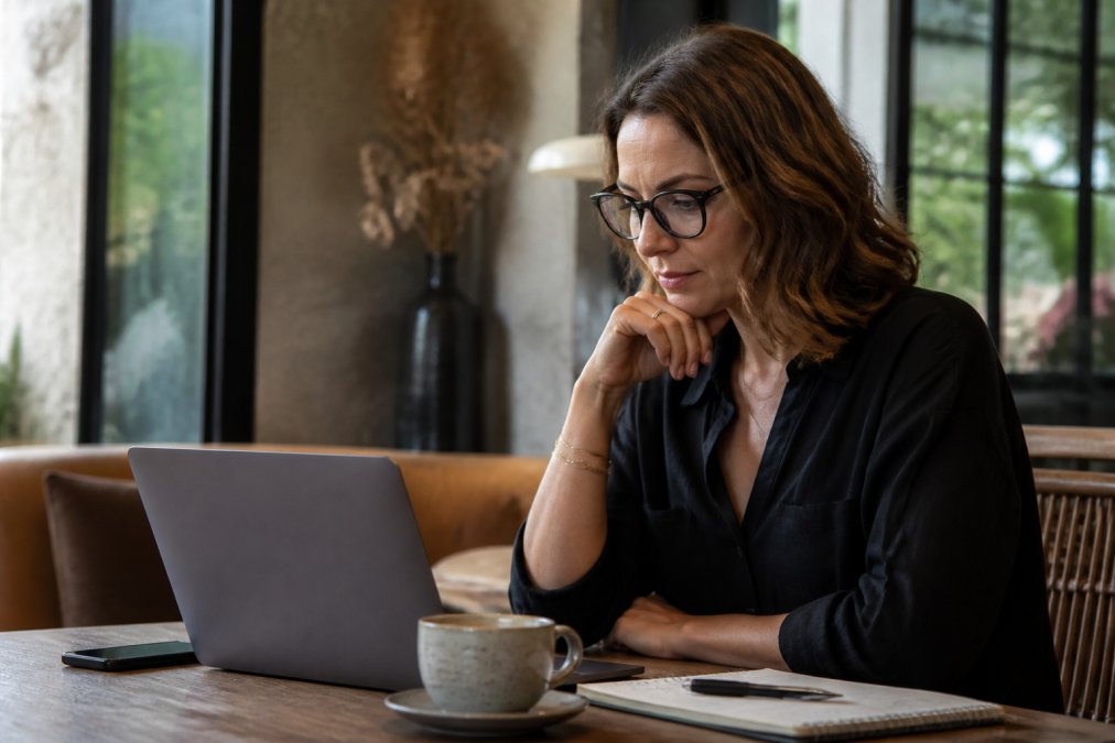 Professional consultant at desk with laptop, notebook, and coffee, reviewing business processes on screen with focused expression in modern office setting