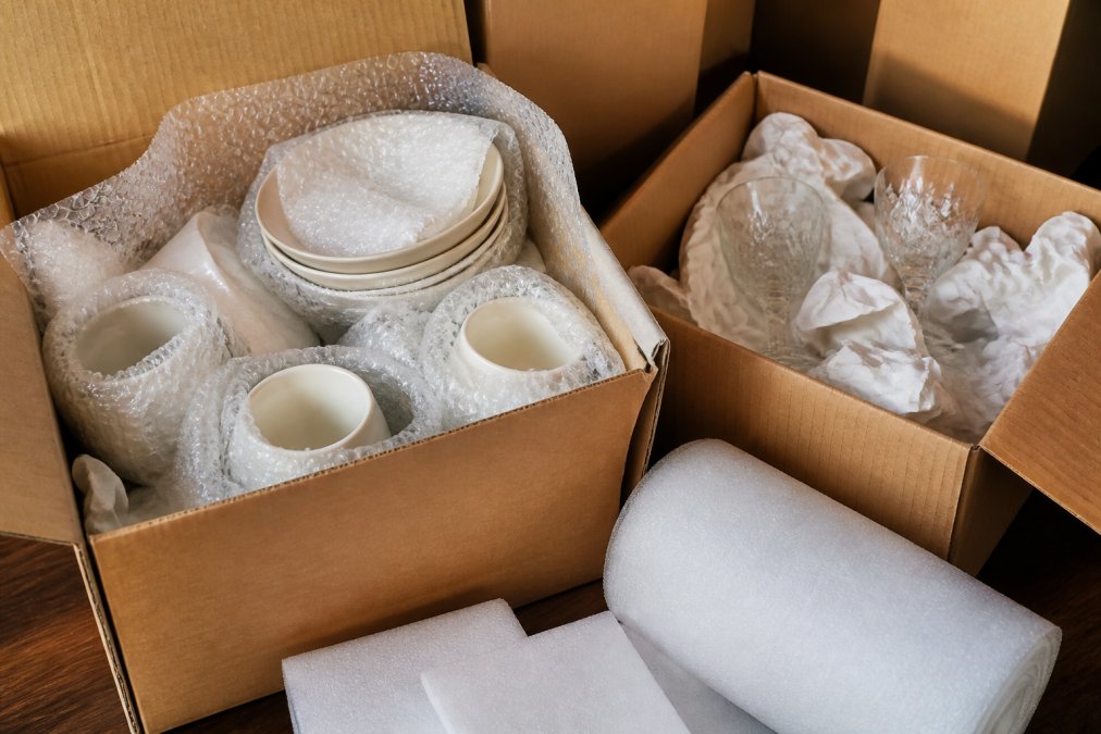 Close-up of carefully wrapped dishes and glassware nestled in bubble wrap and cardboard boxes, showing protective packaging technique with foam sheets visible, organised storage box with tissue paper padding