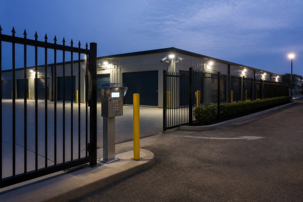 Modern storage facility photographed at dusk with illuminated perimeter lighting, visible CCTV dome cameras mounted on the building corners, and well-lit gate entry system with secure access keypad, security fence surrounding the property